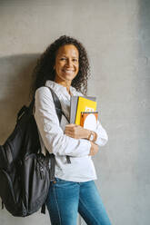 Portrait of smiling woman holding book and backpack leaning on gray wall at college - MASF29265