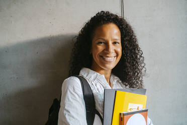 Portrait of smiling woman with curly hair holding book and backpack leaning on gray wall at college - MASF29266