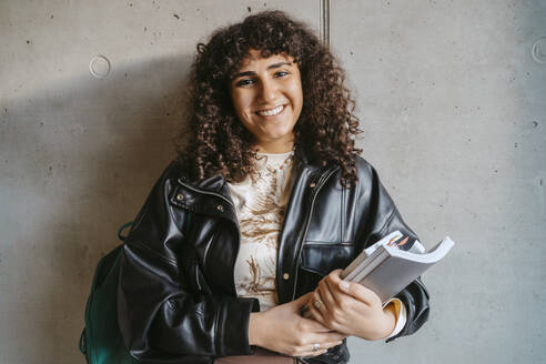 Portrait of smiling young woman holding book and backpack leaning on gray wall - MASF29269