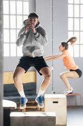 Junger Mann mit Frau macht hüpfende Kniebeugen auf einem Holzkasten im Fitnessstudio - JSRF01932
