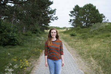 Fröhliche rothaarige Frau steht auf einem Fußweg inmitten einer Wiese - AKLF00528