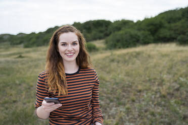 Glückliche junge rothaarige Frau mit Smartphone auf einer Wiese - AKLF00534