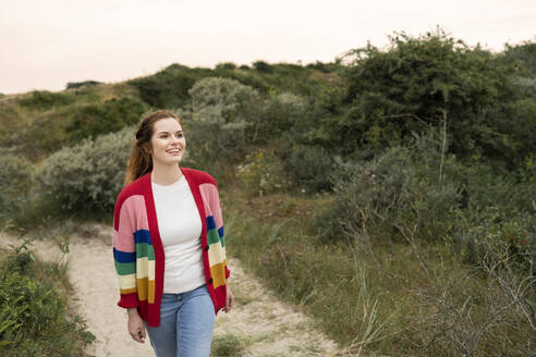 Happy young woman wearing multi colored cardigan walking in meadow - AKLF00540