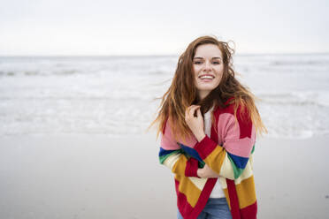 Fröhliche rothaarige Frau in Strickjacke genießt am Strand - AKLF00560