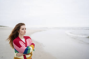 Nachdenkliche junge Frau mit verschränkten Armen am Strand stehend - AKLF00563