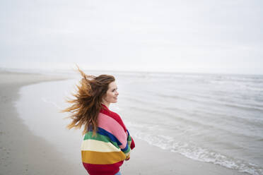 Smiling young woman with tousled hair standing at beach - AKLF00566