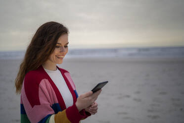 Glückliche junge Frau, die mit ihrem Smartphone am Strand steht und im Internet surft - AKLF00569