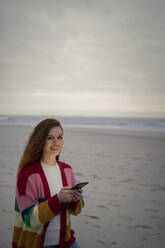 Glückliche schöne junge Frau mit Smartphone am Strand stehend - AKLF00570