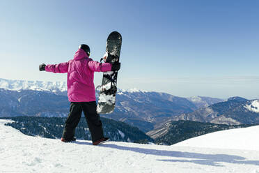Man with arms outstretched holding snowboard in winter on sunny day - OMIF00736