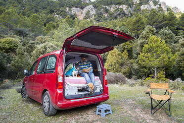 Ganzer Körper eines reifen Mannes, der ein interessantes Buch liest, während er mit seiner französischen Bulldogge in einem Mini-Camper sitzt, während er auf dem Land unterwegs ist - ADSF33876