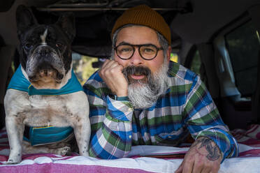 Älterer, bärtiger Mann mit Bart, der in die Kamera schaut, während er mit seiner französischen Bulldogge in einem Mini-Camper auf dem Lande liegt - ADSF33878