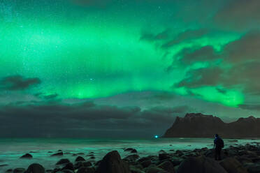 Back view silhouette of anonymous traveler with backpack standing on stony shore and admiring amazing scenery of polar lights in night sky over wavy sea in Lofoten Islands - ADSF33898