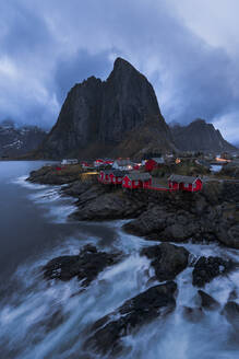 Atemberaubender Blick auf die stürmische See mit schäumendem Wasser und rauen Felsen am Ufer mit kleinen roten Häuschen in Reine, Lofoten in Norwegen - ADSF33908