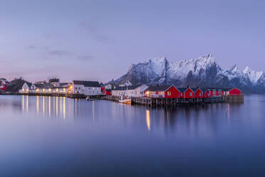 Erstaunlicher Blick auf kleine lebendige Häuser in einem Dorf am Ufer eines ruhigen Flusses gegenüber einem verschneiten hohen Gebirgskamm im Winter abends in Reine, Lofoten Inseln in Norwegen - ADSF33909