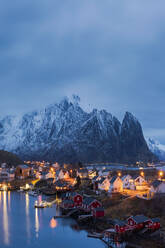 Spectacular view of high snowy mountain range and village with illuminated houses located in bay at bottom of hill in Reine, Lofoten Islands in Norway at sundown - ADSF33912