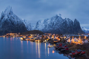 Spektakulärer Blick auf hohe schneebedeckte Berge und ein Dorf mit beleuchteten Häusern in einer Bucht am Fuße eines Hügels in Reine auf den Lofoten in Norwegen bei Sonnenuntergang - ADSF33913