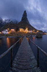 Amazing view of wooden footbridge crossing calm river and leading to illuminated village with cottages located at bottom of highlands in Reine , Lofoten Islands in Norway in evening - ADSF33916