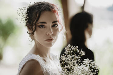 Junge Frau mit Blumenstrauß auf Steinen stehend und in die Kamera schauend in der Nähe eines üppigen Strauches am Hochzeitstag im Garten - ADSF33938