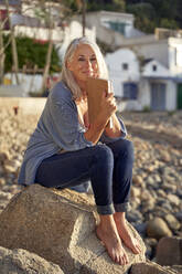 Smiling woman with book sitting on rock at beach - VEGF05399