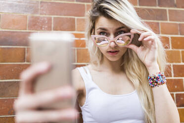 Blondes Teenager-Mädchen mit Sonnenbrille nimmt Selfie durch Smartphone vor der Backsteinmauer - WPEF05830