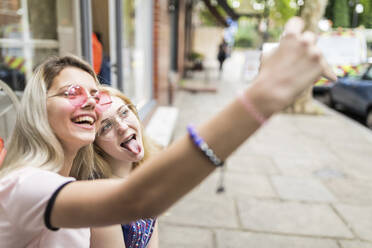Cheerful teenage girl taking selfie through mobile phone with friend sticking out tongue - WPEF05831