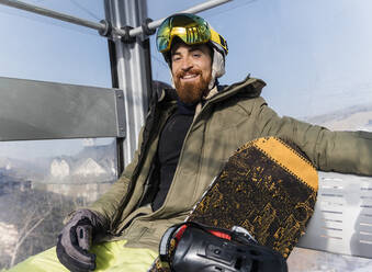 Lächelnder junger Mann mit Snowboard in einer Seilbahn sitzend - JRVF02824