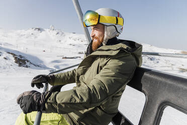 Smiling young man wearing jacket with ski goggles and helmet sitting on ski lift - JRVF02826