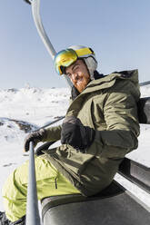 Happy young man gesturing with closed fist sitting on ski lift - JRVF02827