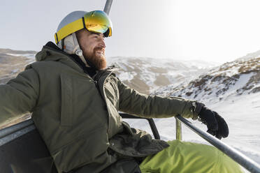 Smiling young man with beard sitting on ski lift - JRVF02830