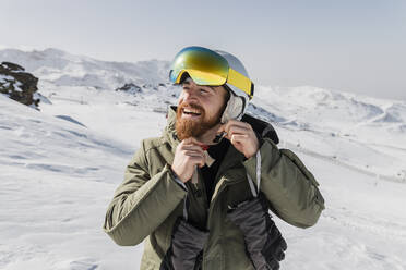 Smiling young man with beard fastening skiing helmet - JRVF02832