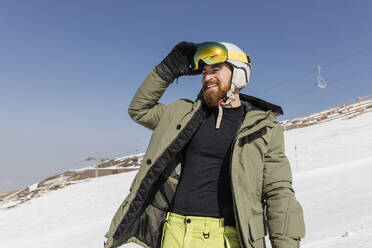 Lächelnder junger Mann mit Skibrille auf einem schneebedeckten Berg an einem sonnigen Tag - JRVF02833