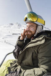 Smiling young man applying suntan lotion sitting on ski lift - JRVF02834