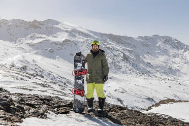 Smiling young man standing with snowboard on rock against snowcapped mountain - JRVF02835