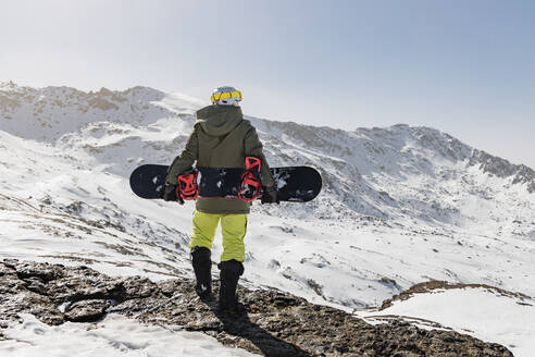 Junger Mann steht mit Snowboard auf einem Felsen - JRVF02837