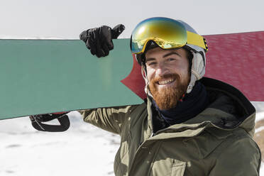 Smiling young man with beard carrying snowboard on shoulder - JRVF02838