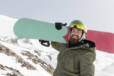 Glücklicher junger Mann mit Bart trägt Snowboard auf der Schulter - JRVF02839