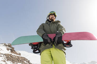 Smiling young man standing with snowboard on sunny day - JRVF02841