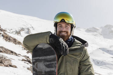 Smiling young man wearing ski goggles standing with snowboard on sunny day - JRVF02844