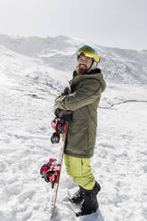 Junger Mann steht mit Snowboard auf einem schneebedeckten Berg - JRVF02845