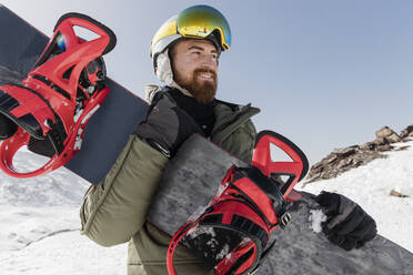 Lächelnder junger Mann mit Skibrille und Snowboard an einem sonnigen Tag - JRVF02846