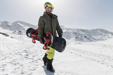 Glücklicher junger Mann mit Snowboard an einem sonnigen Tag - JRVF02850