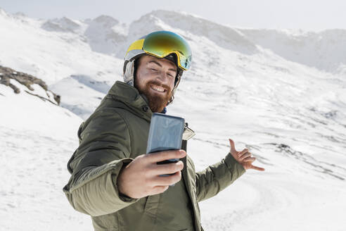 Lächelnder junger Mann gestikuliert bei der Aufnahme eines Selfies durch ein Mobiltelefon an einem sonnigen Tag - JRVF02851