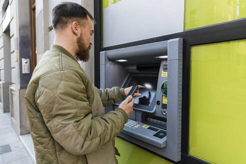 Junger Mann mit Smartphone an einem Geldautomaten - JRVF02858