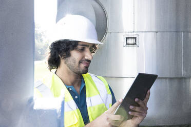 Technician wearing white hardhat holding tablet PC standing in industry - FKF04754