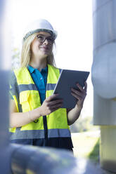 Smiling technician wearing reflective clothing holding tablet PC in industry - FKF04756