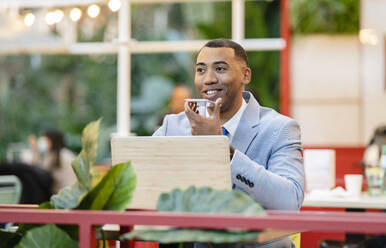 Lächelnder Geschäftsmann, der in einem Straßencafé mit seinem Smartphone über einen Lautsprecher spricht - JCCMF05640