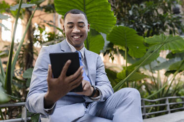 Glücklicher Geschäftsmann mit Tablet-PC vor Pflanzen sitzend - JCCMF05651
