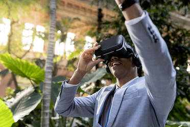 Lächelnder Geschäftsmann mit erhobener Hand im Virtual-Reality-Simulator vor Pflanzen - JCCMF05659