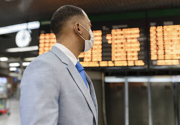 Businessman wearing protective face mask standing in front of departure board at train station - JCCMF05661