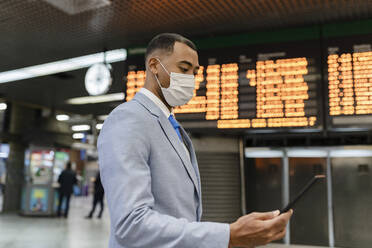 Geschäftsmann mit Gesichtsschutz und Tablet-PC an der Abfahrtstafel eines Bahnhofs - JCCMF05662